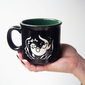 May include: A black ceramic mug with a dark green interior, featuring a white graphic illustration of an otter swimming among seaweed. The mug has a rounded handle and a smooth, glossy finish, showcasing a clean and simple design aesthetic.