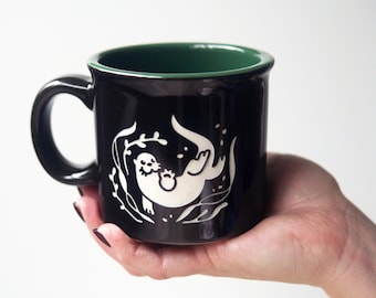 May include: A black ceramic mug with a dark green interior, featuring a white graphic illustration of an otter swimming among seaweed. The mug has a rounded handle and a smooth, glossy finish, showcasing a clean and simple design aesthetic.