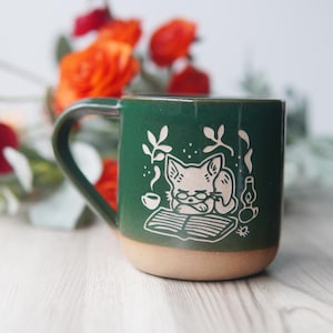 May include: A green ceramic mug with a white illustration of a cat wearing glasses and reading a book. The mug has a brown rim and base.