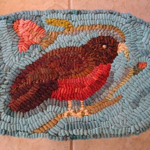May include: A blue and brown rug with a red bird design. The bird is surrounded by a green leaf and a pink flower. The rug is made of yarn and has a textured surface.