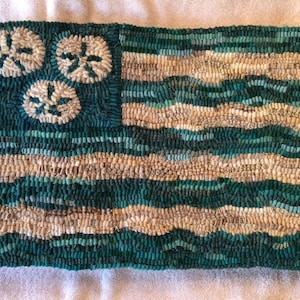 May include: A hand-stitched rug in the shape of an American flag with a blue field and white stars. The stars are in the shape of seashells. The stripes are in shades of blue and white.