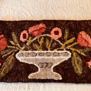 May include: A rectangular, hand-hooked rug featuring a floral design. The rug has a dark brown background with a central vase filled with pink flowers and green foliage. The vase is light gray with a decorative pattern.