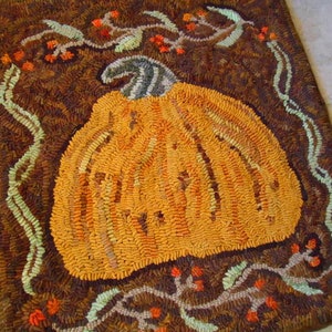 May include: A brown and green rug with a large orange pumpkin in the center. The pumpkin is surrounded by vines and leaves. The rug is made of wool and has a hand-stitched look.