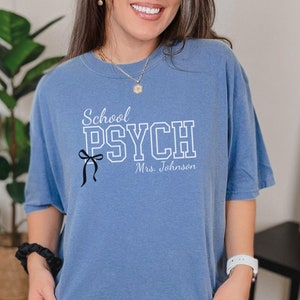 May include: A blue jean t-shirt with the text "School Psych" in white with a black bow and the text "Mrs. Johnson" underneath.