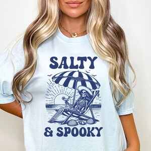 May include: A light blue t-shirt with a graphic of a skeleton sitting in a beach chair under an umbrella with the text "Salty & Spooky" in blue.