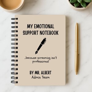 May include: A spiral-bound notebook with the text "MY EMOTIONAL SUPPORT NOTEBOOK" and a pen illustration. The text "...because screaming isn't professional" is below the illustration. A gold pen and a cup of coffee are also visible.