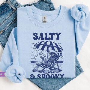 May include: Light blue sweatshirt with a skeleton sitting on a beach chair under an umbrella. The text "Salty & Spooky" is printed on the sweatshirt.
