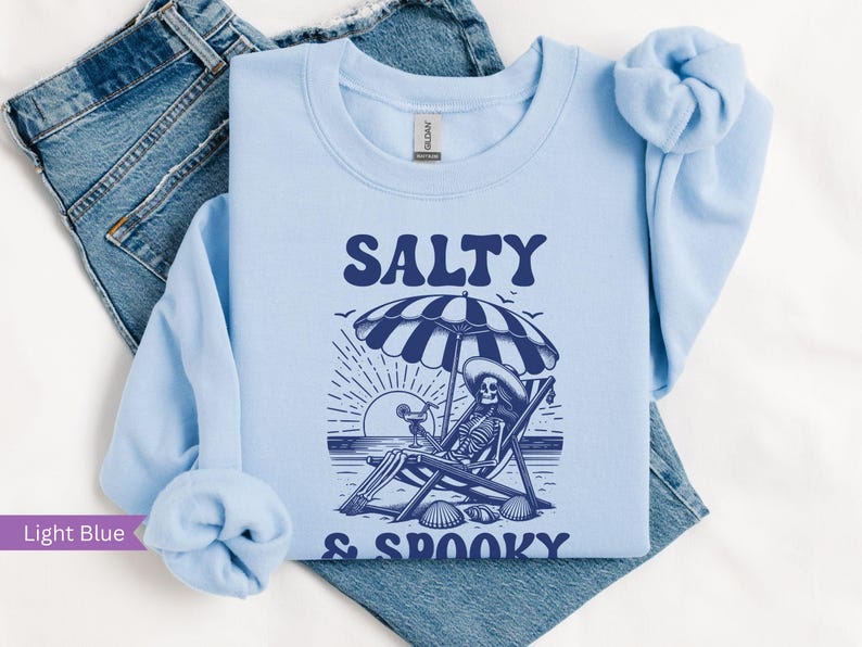 May include: Light blue sweatshirt with a skeleton sitting on a beach chair under an umbrella. The text "Salty & Spooky" is printed on the sweatshirt.