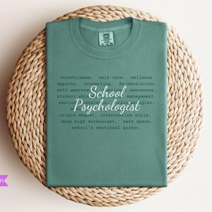 May include: A light green t-shirt with the text "School Psychologist" defined as "mindfulness, self-care, wellness, empathy, counseling, de-escalation, self-awareness, emotion awareness, student whisperer, brain wrangler, emotion translator, crisis magnet, intervention ninja, deep sigh enthusiast, safe space, school's emotional anchor."