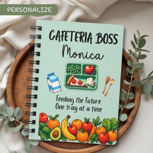 May include: A spiral-bound notebook with a light green cover featuring the text "CAFETERIA BOSS Monica." The notebook has an illustration of a school lunch tray with peas, an apple, pizza, and a carton of milk. The bottom border has illustrations of fruits and vegetables.