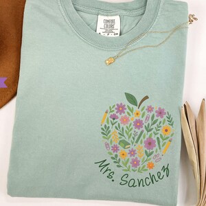 May include: A light green t-shirt featuring a floral apple design with pencils, flowers, and the text "Mrs. Sanchez". A gold necklace rests on the shirt. The shirt is made of a soft material.