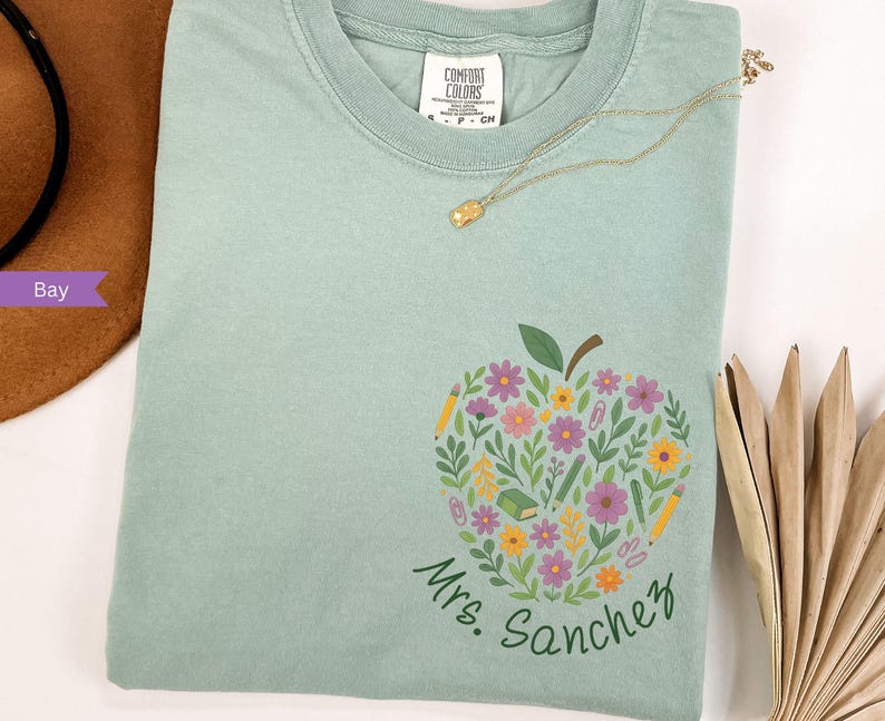 May include: A light green t-shirt featuring a floral apple design with pencils, flowers, and the text "Mrs. Sanchez". A gold necklace rests on the shirt. The shirt is made of a soft material.