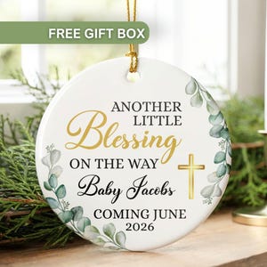 May include: A white ceramic ornament with gold text and a cross, featuring the words "ANOTHER LITTLE Blessing ON THE WAY Baby Jacobs COMING JUNE 2026." The ornament is decorated with green and gray leaves and has a gold string for hanging.