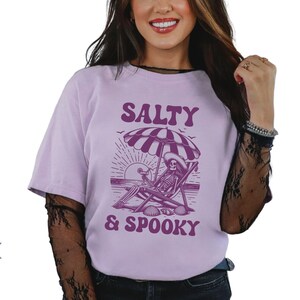 May include: A purple t-shirt with a graphic of a skeleton sitting in a beach chair under an umbrella. The text "Salty & Spooky" is printed on the shirt.