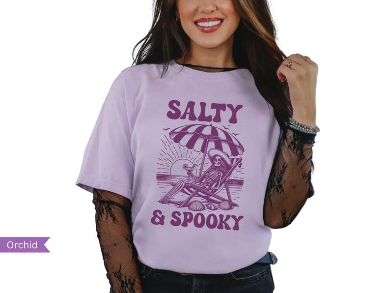 May include: A purple t-shirt with a graphic of a skeleton sitting in a beach chair under an umbrella. The text "Salty & Spooky" is printed on the shirt.