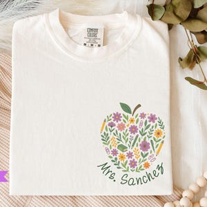 May include: Ivory-colored t-shirt with a floral apple design. The apple is filled with purple, yellow, and white flowers, green leaves, and school supplies. The name "Mrs. Sanchez" is written in green script below the apple.