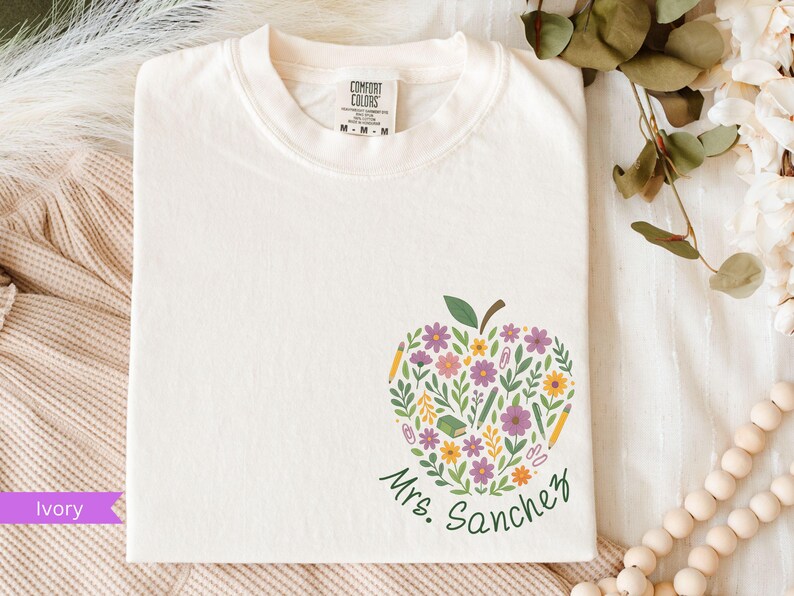 May include: Ivory-colored t-shirt with a floral apple design. The apple is filled with purple, yellow, and white flowers, green leaves, and school supplies. The name "Mrs. Sanchez" is written in green script below the apple.