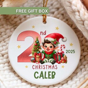 May include: A white ceramic Christmas ornament with a festive design. It features a cartoon elf, a Christmas tree, and the text "2nd Christmas Caleb 2025". The ornament has a gold string for hanging.