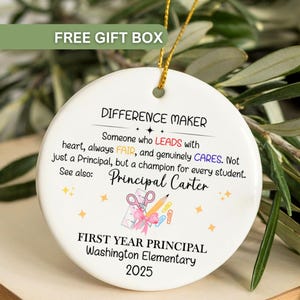 May include: A white ceramic ornament with gold string, featuring text that reads "DIFFERENCE MAKER" and "FIRST YEAR PRINCIPAL". The ornament includes a graphic of school supplies and is personalized with a name and the year 2025.