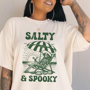 May include: A white t-shirt with a green graphic design of a skeleton sitting in a beach chair under an umbrella. The text "Salty & Spooky" is printed below the image.