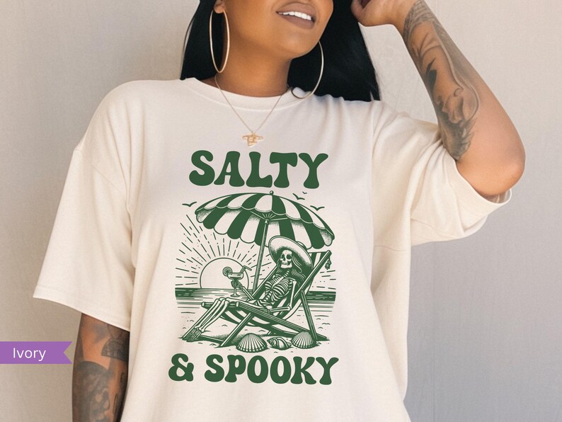 May include: A white t-shirt with a green graphic design of a skeleton sitting in a beach chair under an umbrella. The text "Salty & Spooky" is printed below the image.