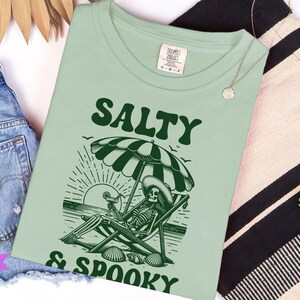 May include: A green t-shirt with a skeleton sitting in a beach chair under an umbrella. The text "Salty & Spooky" is printed on the shirt.