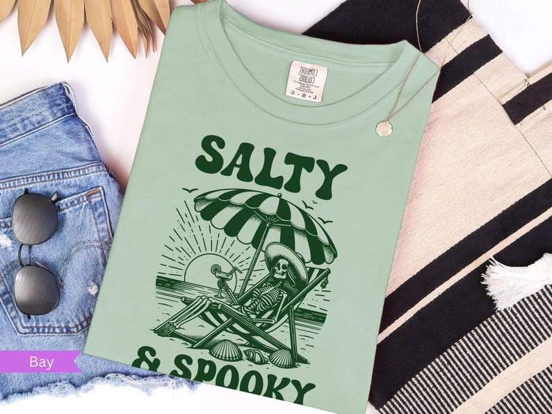 May include: A green t-shirt with a skeleton sitting in a beach chair under an umbrella. The text "Salty & Spooky" is printed on the shirt.