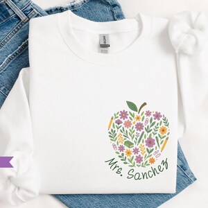 May include: White long-sleeve sweatshirt with a floral apple design. The apple is filled with flowers, pencils, and a book. The name "Mrs. Sanchez" is written in green script below the apple.