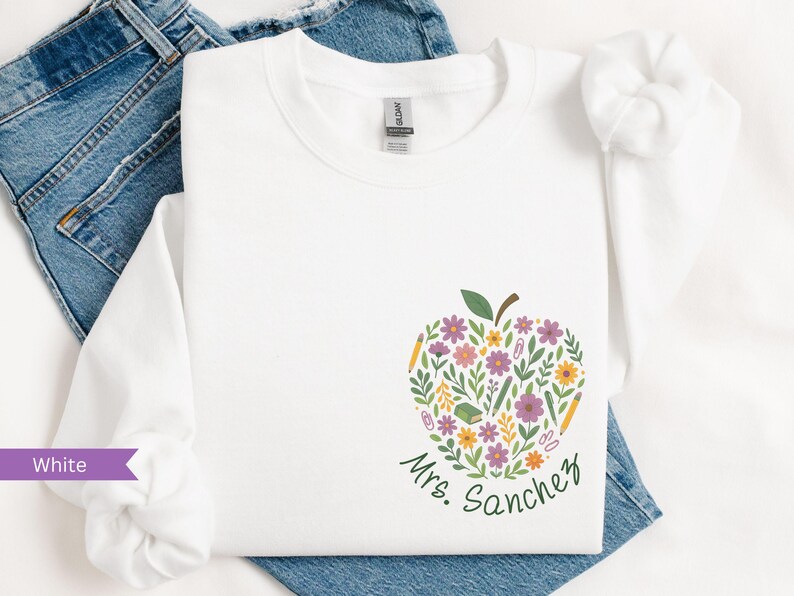 May include: White long-sleeve sweatshirt with a floral apple design. The apple is filled with flowers, pencils, and a book. The name "Mrs. Sanchez" is written in green script below the apple.