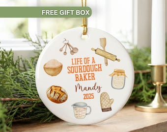 May include: A white ceramic ornament with a gold string, featuring watercolor illustrations of baking tools and bread. Text reads "Life of a Sourdough Baker" with a name and the year 2025. The ornament is a gift.