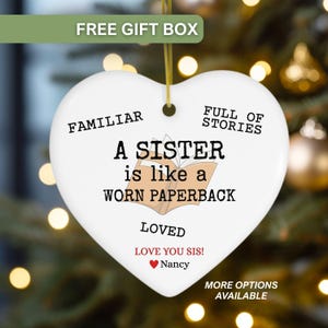 May include: White heart-shaped ceramic ornament with gold string, featuring text: "A SISTER is like a WORN PAPERBACK." Additional text includes "FAMILIAR," "FULL OF STORIES," "LOVED," and "LOVE YOU SIS! Nancy." The ornament is for a sister.