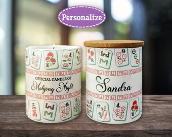May include: Two white ceramic candles with Mahjong tile designs. One candle is lit, and the other has a wooden lid. Text on the candles reads "Official Candle of Mahjong Night" and "Sandra".