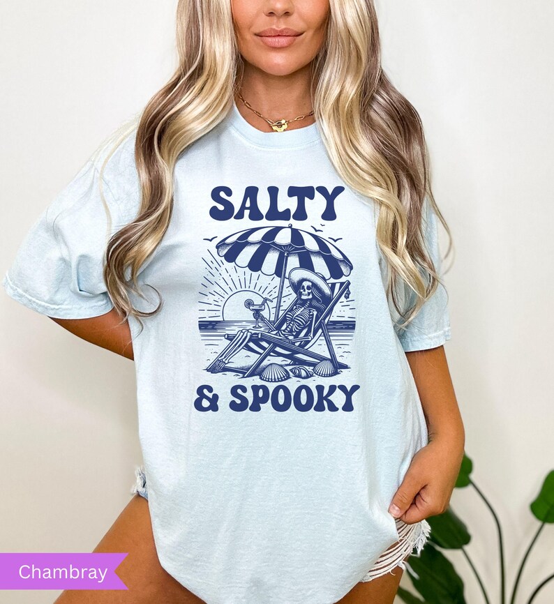 May include: A light blue t-shirt with a graphic of a skeleton sitting in a beach chair under an umbrella with the text "Salty & Spooky" in blue.