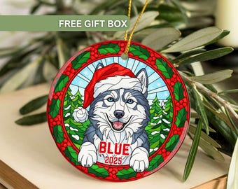 May include: A round Christmas ornament featuring a Siberian Husky wearing a Santa hat. The ornament has a stained-glass design with a holly and pine tree border. The word "BLUE" and the year "2025" are displayed.