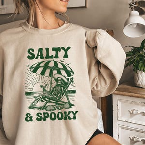 May include: A beige sweatshirt with a green graphic of a skeleton sitting on a beach chair under an umbrella. The text "Salty & Spooky" is printed on the sweatshirt.