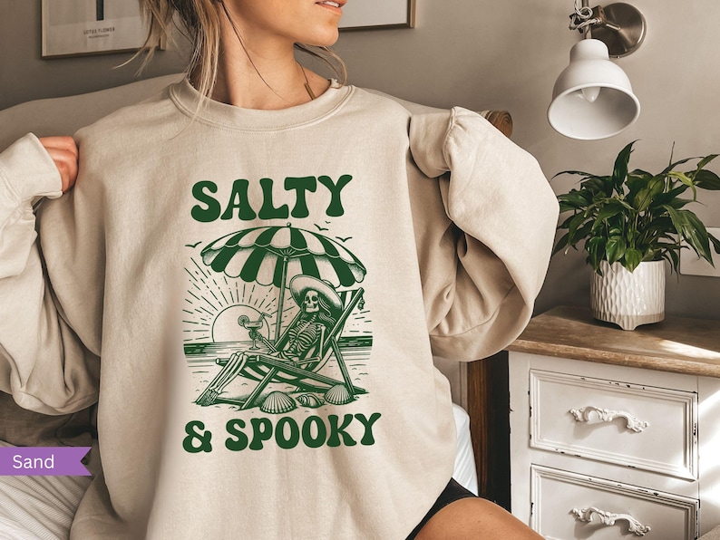 May include: A beige sweatshirt with a green graphic of a skeleton sitting on a beach chair under an umbrella. The text "Salty & Spooky" is printed on the sweatshirt.