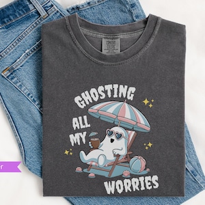 May include: A dark gray t-shirt with a graphic of a ghost wearing sunglasses and relaxing on a beach chair under an umbrella. The text "Ghosting all my worries" is printed on the shirt.
