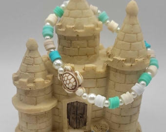 May include: A beaded bracelet featuring a central turtle charm, alternating with white faux pearls, turquoise blue disc beads, and cream-coloured disc beads. The bracelet is strung on an elastic cord, creating a circular shape with a repeating pattern of beads.