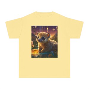 Op de afbeelding: Een lichtgeel t-shirt met een centrale grafische print van een klein, harig, lichtbruin wezen met grote donkere ogen, zittend bij een gloeiende honingraat in een kleurrijk landschap met bloemen en een zonsondergang op de achtergrond.