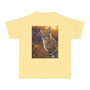 Puede incluir: Una camiseta de manga corta de color amarillo pálido con un estampado gráfico rectangular centrado de un serval de pie en un campo con flores silvestres moradas al atardecer. El felino tiene pelaje beige con manchas negras y orejas grandes.