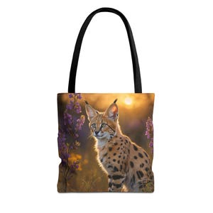 Puede incluir: Un bolso tote con una impresión fotográfica de un serval de pelaje moteado sentado entre flores silvestres moradas al atardecer. El bolso cuenta con dos asas de tela negra sólida fijadas en la parte superior para facilitar su transporte.