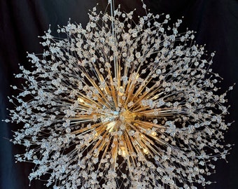 May include: A large, round, crystal chandelier with a gold finish. The chandelier has many arms that extend outward, each with a crystal bead at the end. The chandelier is hanging from a ceiling.