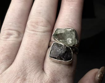 May include: A silver ring with two raw gemstones. The top stone is a light green, and the bottom stone is a dark purple.