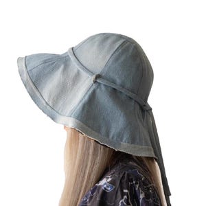 May include: A light blue denim sun hat with a wide brim and a tie detail. The hat has a frayed edge and a soft, worn appearance. The hat is designed to provide sun protection and is a stylish accessory.