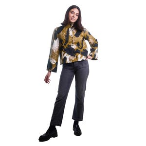 May include: A woman wearing a button-up shirt with a unique abstract pattern in shades of olive green, black, and white. She is also wearing dark gray jeans and black shoes. The shirt has long sleeves and a high collar.