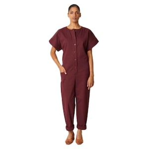 May include: A burgundy-colored jumpsuit with short sleeves and a button-up front. The jumpsuit has a relaxed fit with pockets and cuffed ankles. The model is wearing brown shoes. The overall style is casual and comfortable.