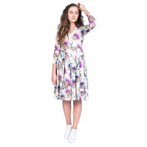 May include: A white dress with a floral print of purple and pink thistles. The dress has long sleeves and a pleated skirt. The model is wearing white sneakers.