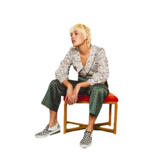 May include: A person wearing a white and floral patterned blouse, green and black patterned pants, and black and white checkered slip-on shoes. They are sitting on a red cushioned stool with a wooden frame.