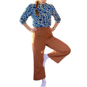May include: A blue and white floral print blouse with three-quarter sleeves and a high neckline. The blouse is paired with high-waisted, wide-leg, rust-colored pants. The model is wearing white socks.
