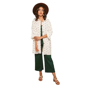 May include: A white long-sleeved shirt with a red polka dot pattern, worn over a dark green jumpsuit. The person is wearing a brown hat and brown sandals.